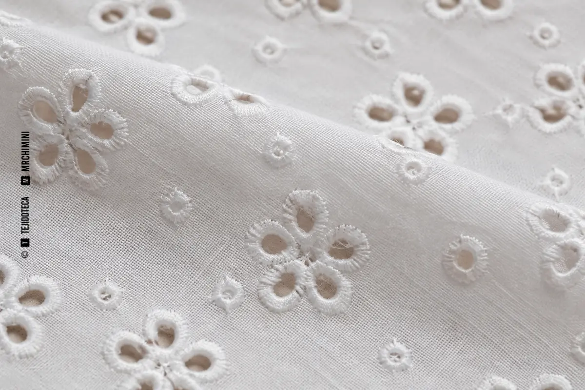 Detalle macro de algodón blanco con ojetes florales rodeados por bordado inglés.