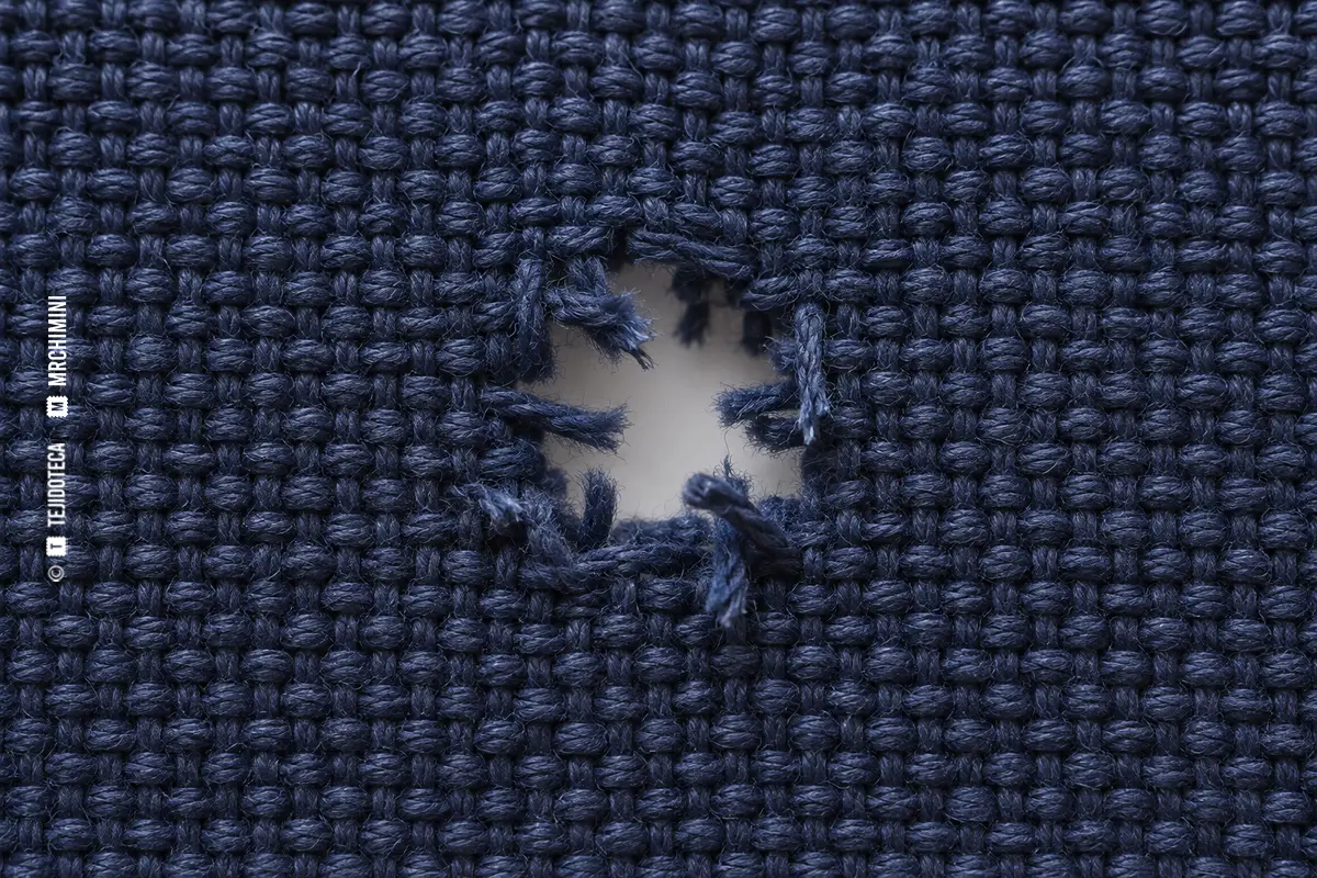 Macro de tejido azul con pequeño agujero y extremos rotos de urdimbre y trama