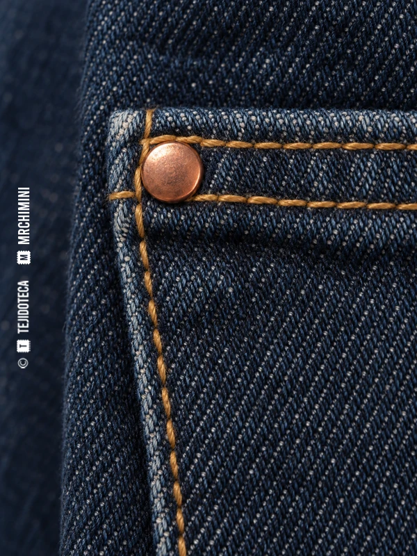 Detalle de un bolsillo de denim índigo con pespuntes y un remache cobrizo en la esquina.